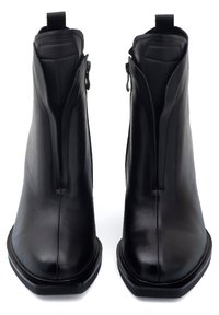 Stivali alla caviglia in pelle nera con punta affusolata, cerniere laterali e suola piatta e squadrata. Texture liscia e design minimalista.