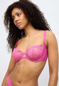 Ροζ μπραλέτα από δαντέλα με κυματιστή άκρη, διάφανα κύπελλα και διακοσμητικό εξάρτημα στο κέντρο. Οι ρυθμιζόμενες τιράντες ενισχύουν την εφαρμογή και τη στήριξη.