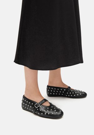 Czarne skórzane baleriny z okrągłym noskiem, ozdobione srebrnymi ćwiekami i dekoracyjnym paskiem z klamrą, osadzone na czarnej spódnicy.