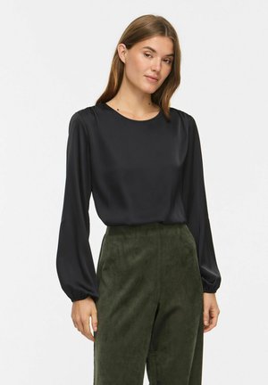 Femme portant un chemisier noir à manches longues et un pantalon vert foncé taille haute, debout devant un fond blanc.