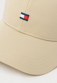 Béžová baseballová čepice vyrobená z texturované látky, s malou nášivkou loga v námořnické modré, červené a bílé barvě na přední straně. Lehce zakřivený kšilt.