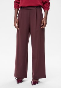 Bordowe spodnie o szerokich nogawkach z gładką fakturą, z przodu plisowania i dwoma ciemnymi guzikami na pasku. Noszone z błyszczącymi, szpiczastymi butami.