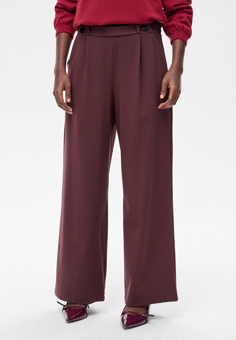 Bordowe spodnie o szerokich nogawkach z gładką fakturą, z przodu plisowania i dwoma ciemnymi guzikami na pasku. Noszone z błyszczącymi, szpiczastymi butami.