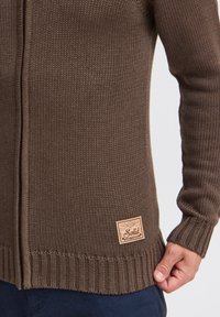 Cardigan lavorato a maglia marrone con bordo e polsini a coste. Presenta una toppa in pelle con la scritta "Solid" sul lato sinistro in basso.