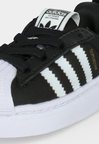 Αθλητικό παπούτσι Adidas Superstar ασπρόμαυρο με μαύρα κορδόνια, λευκή μύτη, τρεις λευκές ρίγες και χρυσό κείμενο "Superstar" στο πλάι.