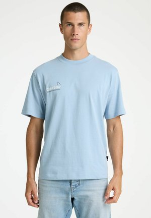 Rövid hajú férfi világoskék pólóban, melyen fehér "CHASIN'" felirat látható, és világoskék farmerben, egyszerű fehér háttér előtt áll.