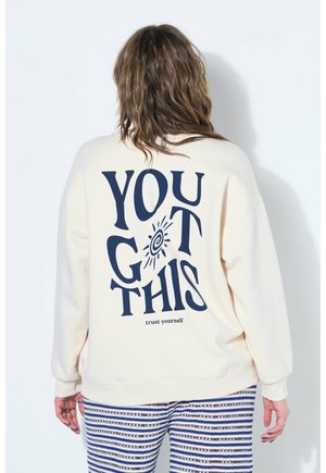 Person med bølget hår iført en cremefarvet sweatshirt med teksten "YOU GOT THIS" og solgrafik, kombineret med blå og hvide stribede bukser.