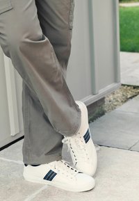 Weiße Sneaker mit blauen und grauen Streifen, runde Spitze, Schnürdesign, getragen mit hellgrauen Cargo-Hosen auf einer gepflasterten Fläche.
