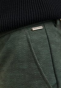 Tessuto verde a zigzag con una superficie texture, caratterizzato da un'etichetta argentata "JACK & JONES" su un maglione di lana scura sopra.