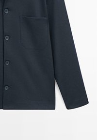 Giacca blu navy scuro con bottoni, una tasca sul petto e maniche lunghe su sfondo bianco.