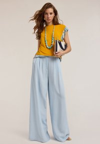 Geel top met bloemenrand, gecombineerd met lichtblauwe wijde broek. Gemaakt met een blauwe kralenketting en een kleine handtas.