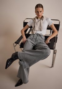 Tenue décontractée comprenant un t-shirt blanc ajusté et un jean large en denim gris. Bottines noires à talons avec des clous argentés. Pull léger autour du cou.