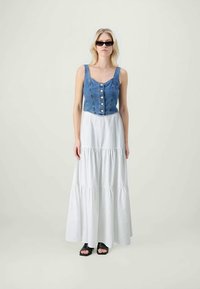 Denimová croptop s knoflíky, zkombinovaná s dlouhou bílou sukní s volány. Model nosí černé sluneční brýle a černé sandály na otevřenou špičku.