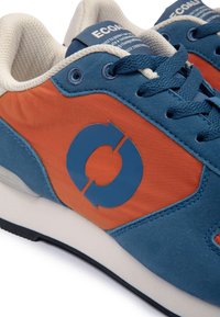 Baskets bleues et orange avec une doublure crème, des lacets bleus et un logo flèche circulaire sur le côté, à côté de la semelle et des œillets.
