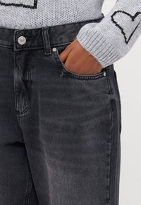Crne traperice od denim s prednjim džepom i srebrnim kopčanjem. Tkanina ima glatku teksturu i suptilne detalje izbljeđenja.