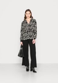 Blusa preta com estampado floral, decote em V e mangas longas, combinada com calças largas pretas. A modelo segura uma bolsa preta e usa botas de tornozelo.