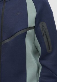 Sweatshirt com capuz azul-marinho e verde-azulado, feito de tecido suave, apresentando um logo preto, texturas nas laterais e um bolso com fecho no braço.