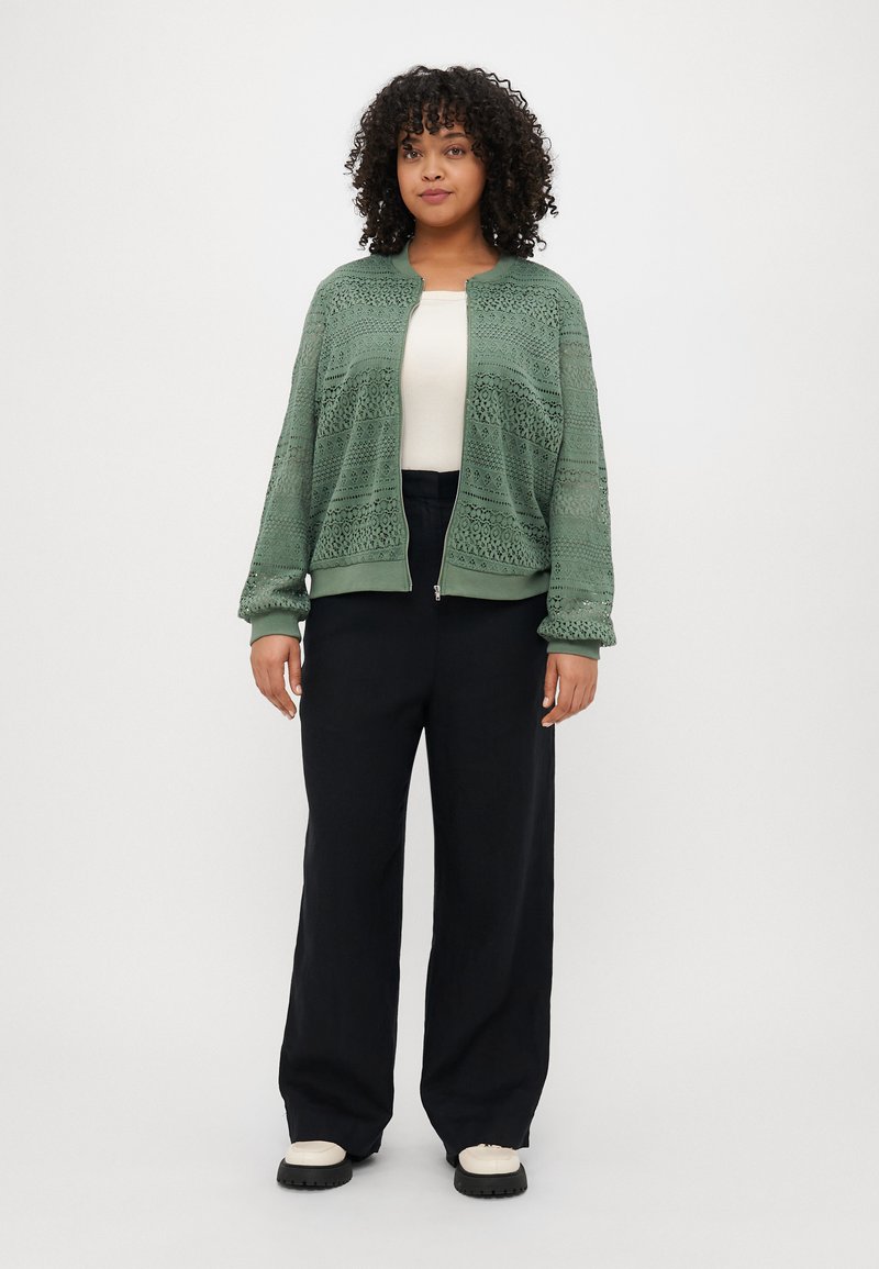 Zielona, lekka dzianinowa kurtka z misternym wzorem koronkowym, ze ściągaczami przy rękawach i u dołu. Nosi ją osoba w białej bluzce oraz czarnych szerokich spodniach i butach.