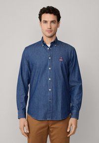 Donkerblauwe denimshirt, lange mouwen en met een knoopsluiting aan de kraag. Heeft een geborduurd logo op de linker borst en witte knopen. Glad van textuur.