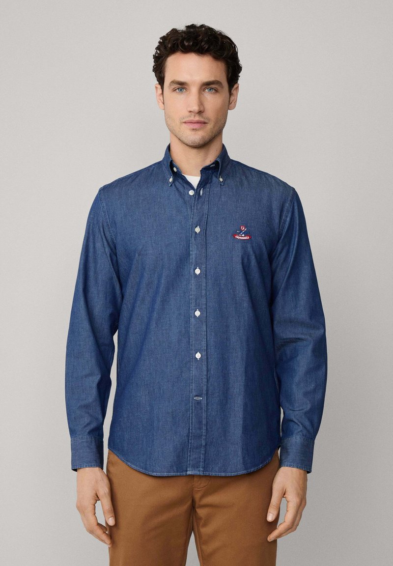 Donkerblauwe denimshirt, lange mouwen en met een knoopsluiting aan de kraag. Heeft een geborduurd logo op de linker borst en witte knopen. Glad van textuur.