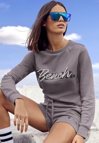 Grijze sweatshirt met het logo "Bench.", gecombineerd met bijpassende shorts. Model draagt blauwe spiegeltjeszonnebril en enkelsokken met zwarte strepen.