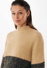 Suéter de punto beige con un cuello acanalado alto. La sección inferior presenta un patrón de espiga negro. Textura suave, corte holgado.