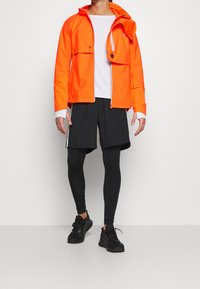 Giacca zip arancione brillante con due tasche frontali, abbinata a pantaloni corti neri e leggings neri. Scarpe da ginnastica nere completano il look atletico.