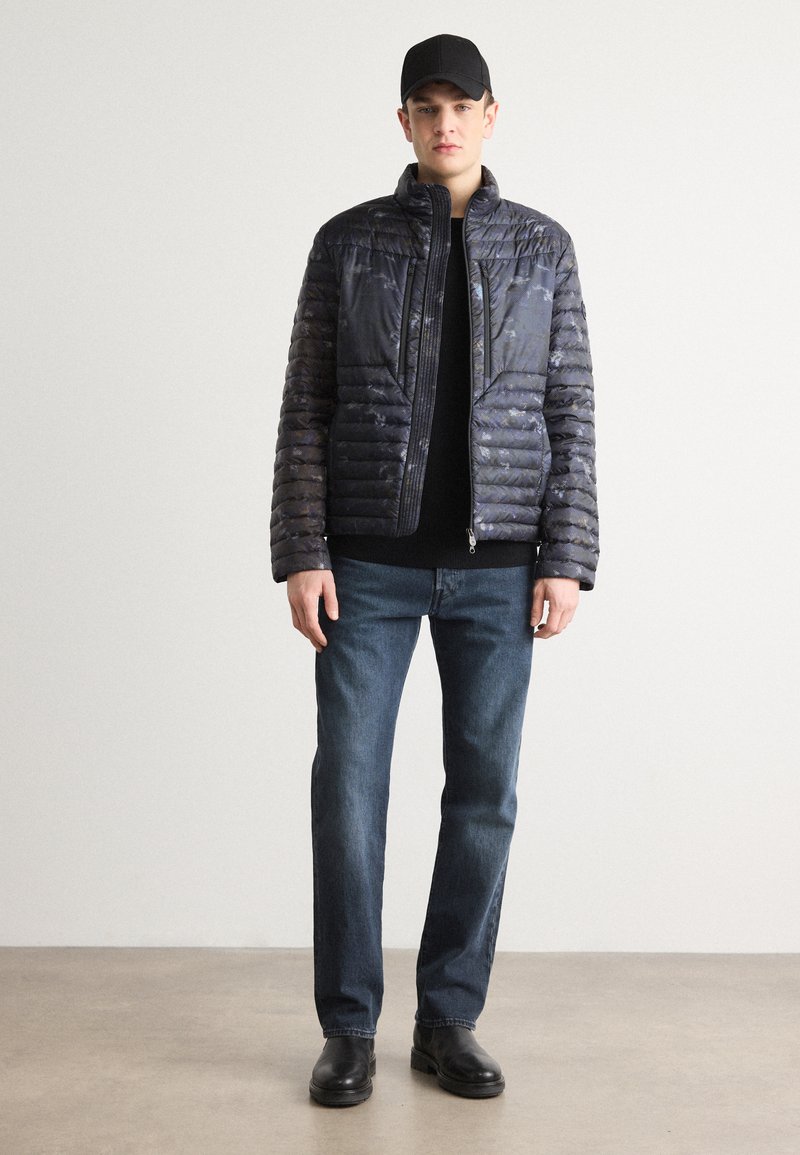 Czarna, pikowana kurtka puchowa z wysokim kołnierzem i pełnym zamkiem. Noszona na czarnym swetrze, w połączeniu z niebieskimi dżinsami i czarnymi butami.