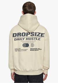 Kremasti dukserica s velikim crnim grafičkim natpisom na leđima. Ima kapuljaču s vezicom i rebraste manžete. Materijal izgleda mekan i teksturiran.