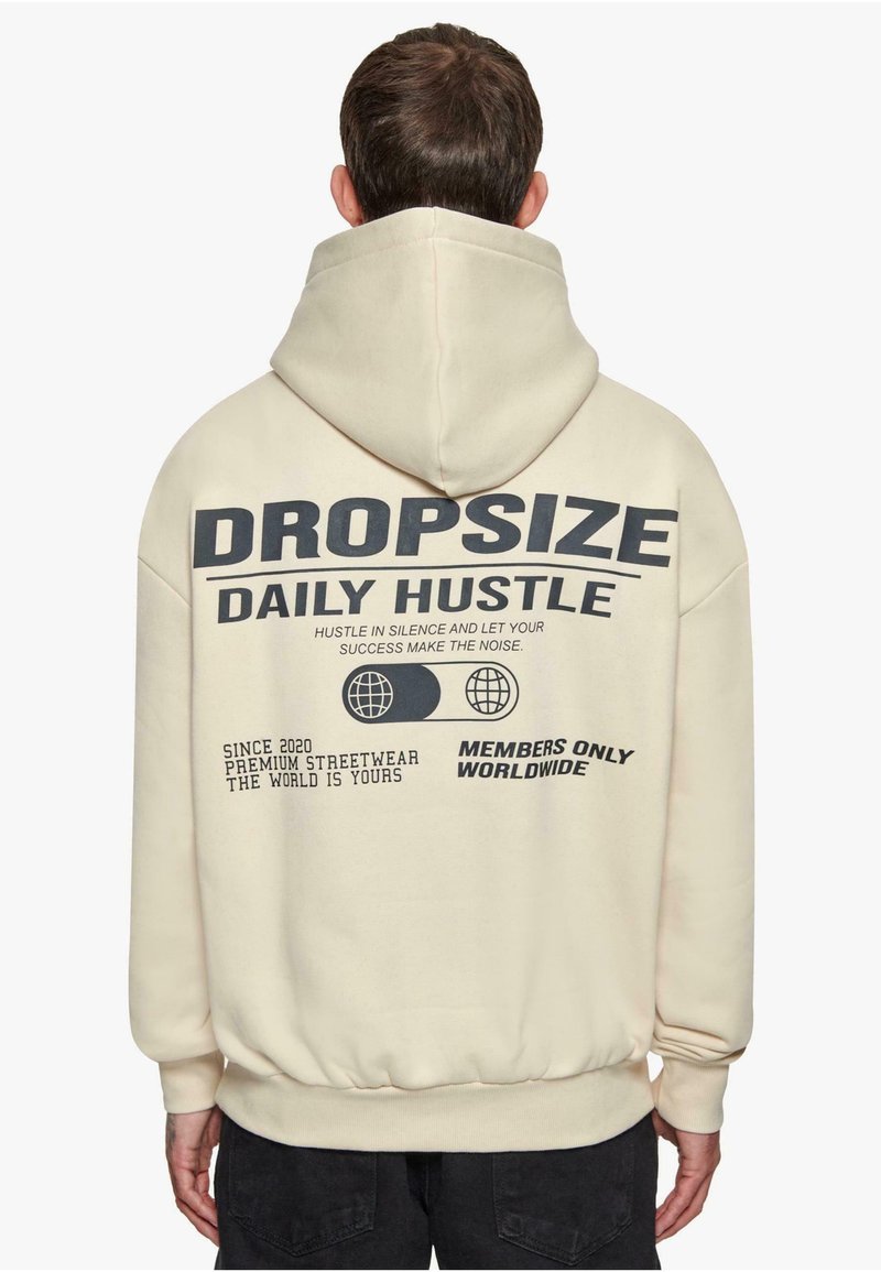 Kremasti dukserica s velikim crnim grafičkim natpisom na leđima. Ima kapuljaču s vezicom i rebraste manžete. Materijal izgleda mekan i teksturiran.