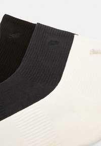 Tre par ribbede sokker i sort, mørkegrå og hvid med subtil branding. Hver sok har en ensfarvet nuance og en blød tekstur.
