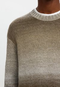 Plano detalle de una persona que lleva un suéter de punto degradado beige y marrón con cuello redondo acanalado sobre una camisa blanca.