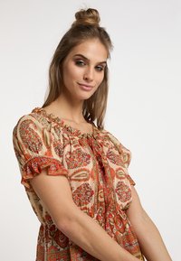 Blusa a stampa floreale in beige con accenti rossi. Presenta un colletto raggruppato, maniche corte con volant e una vestibilità rilassata. Tessuto con una consistenza liscia.