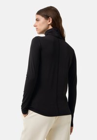 Vrouw met bruin haar, gekleed in een zwart langslijfig coltrui en crèmekleurige broek, staand met handen in de zakken, met de gezicht naar links gericht.