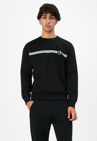 Young man with dark hair wearing a black sweatshirt with white stripes and symbols across the chest, standing with one hand in pocket.