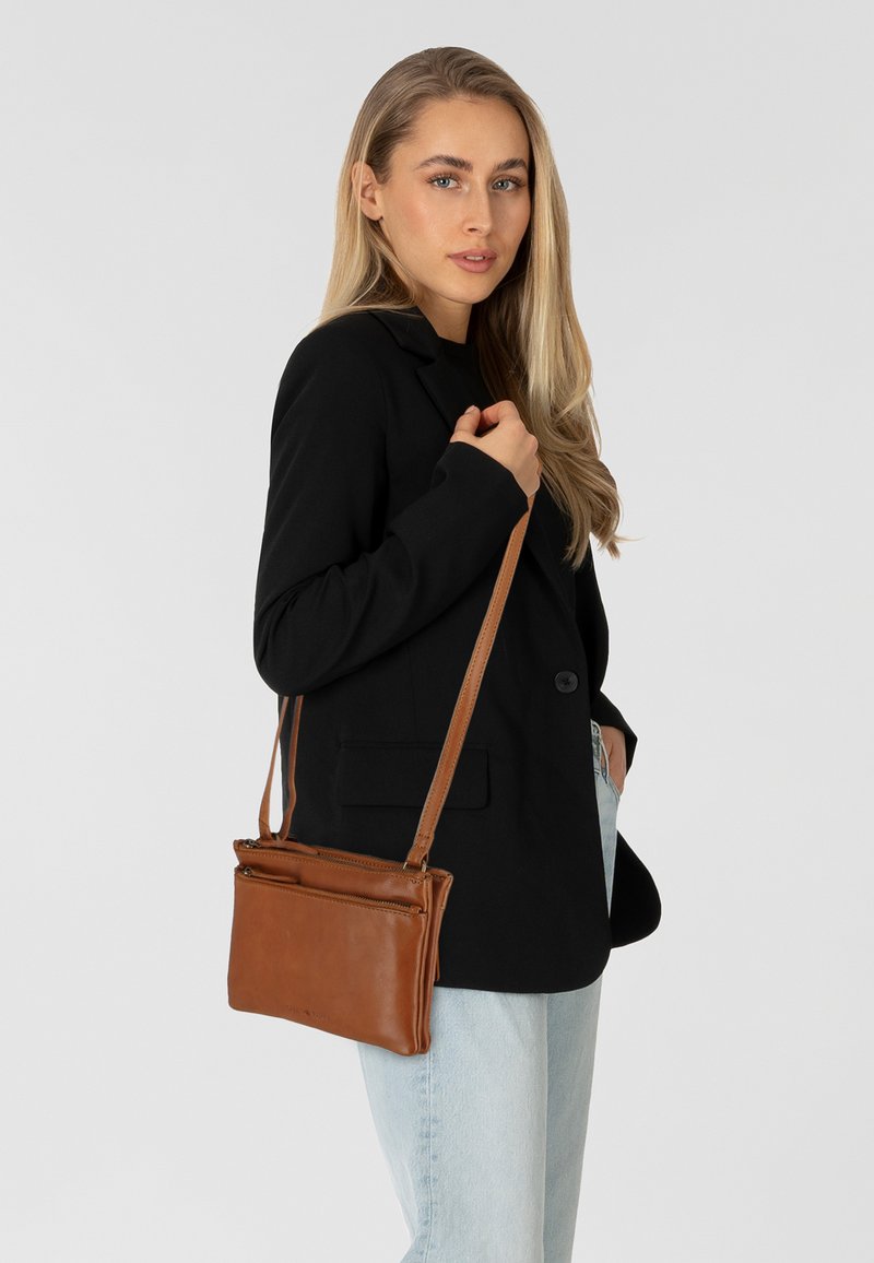Borsa a tracolla in pelle marrone con due tasche con zip, design sottile e tracolla staccabile, indossata sopra un blazer nero.