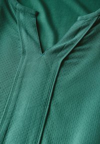 Tessuto verde scuro con un motivo testurizzato e perforato. Scollo a V e dettagli di cucitura visibili accentuano il design del capo.