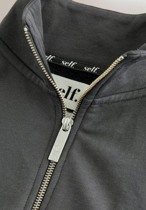 Hallatav hall tõmblukuga jakk lähedalt, millel on metallist tõmblukk, kõrge krae ja sile tekstuur; taga on silt, mis kuvab sõna "self." kontrastsetes kirjades.