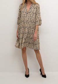 Zwart- en beigegekleurd jurkje met patroon, diepe V-hals, driekwart mouwen en een vloeiende silhouet, gecombineerd met zwarte hoge hakken.