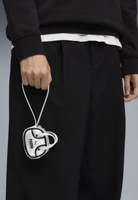 Zaino mini bianco con accenti argentati e manico a coulisse, tenuto in una mano. Indossato con un outfit nero, caratterizzato da pantaloni plissettati.