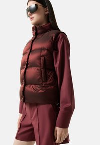 Bordeauxroter Puffer-Weste mit hohem Kragen, ausgestattet mit horizontalen Nähten, einer Vordertasche, und getragen über einem passenden Hemd und Shorts.