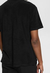 Personne portant une chemise noire à manches courtes et un pantalon noir, vue de dos sur un fond clair et uni.