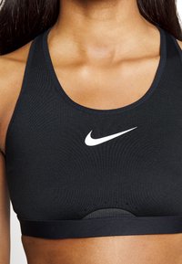 Μαύρο αθλητικό σουτιέν με στρογγυλή λαιμόκοψη και λογότυπο Nike. Διαθέτει υφή υλικού και διάτρητες λεπτομέρειες για καλύτερη αναπνοή.