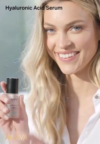 Przezroczysta szklana butelka serum z kwasem hialuronowym z czarną nakrętką, oznaczona etykietą "AHAVA". Modelka trzyma butelkę, pokazując jej zawartość, w delikatnym świetle.