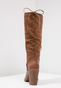 Botte marron montant jusqu'au genou avec une tige en daim souple et un talon en cuir texturé. Dispose d'un lien décoratif en haut et d'une fermeture éclair latérale.