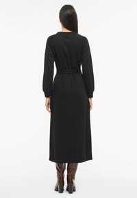 Černé pletené šaty s dlouhým rukávem, s páskem v pase a volným střihem, se zaobleným výstřihem a žebrovanou strukturou. Délka sahá po půl lýtka.