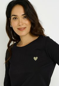 Top negro de manga larga con cuello redondo que presenta un pequeño diseño de corazón dorado en el pecho, hecho de tela suave y elástica.