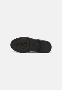 Suela de zapato negra con superficie texturizada; presenta un patrón circular de agujeros y una impresión de logo. Material de goma, perfil plano.
