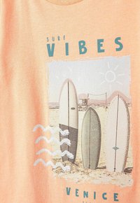 Meruňkové tričko s grafikou surfování obsahující tři surfovací prkna, modrý text "SURF VIBES" a vlnité bílé akcenty.