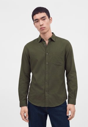 Homme portant une chemise olive à manches longues boutonnée et un jean bleu foncé, debout devant un fond clair uni.